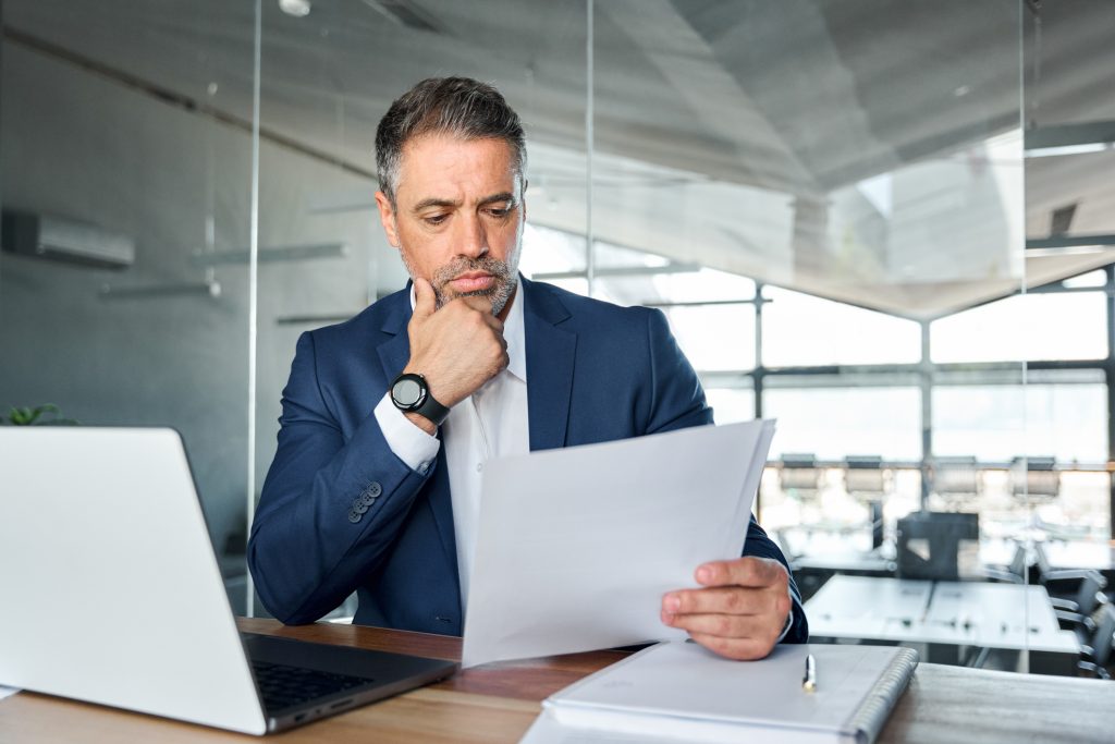 Serious busy mid aged professional business man lawyer or financial law expert wearing suit holding corporate documents reading paper contract sitting at desk in office managing risks feeling doubt.
